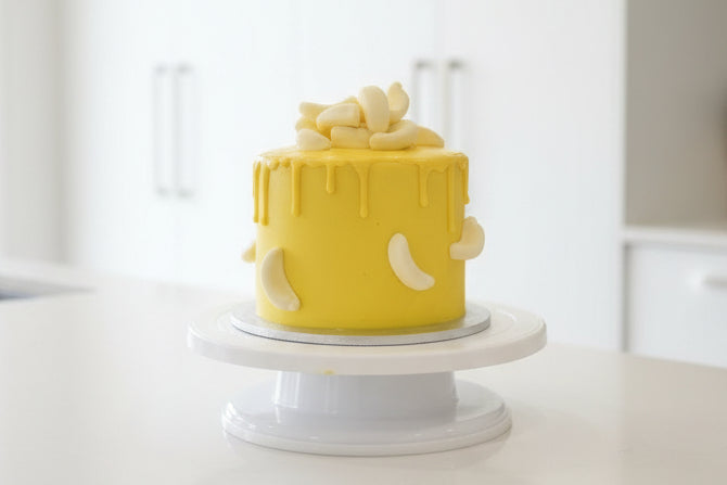 A yellow frosted cake with yellow drip and banana lollies on top, displayed on a white cake stand on a dark kitchen countertop.