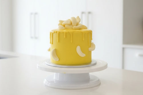A yellow frosted cake with yellow drip and banana lollies on top, displayed on a white cake stand on a dark kitchen countertop.