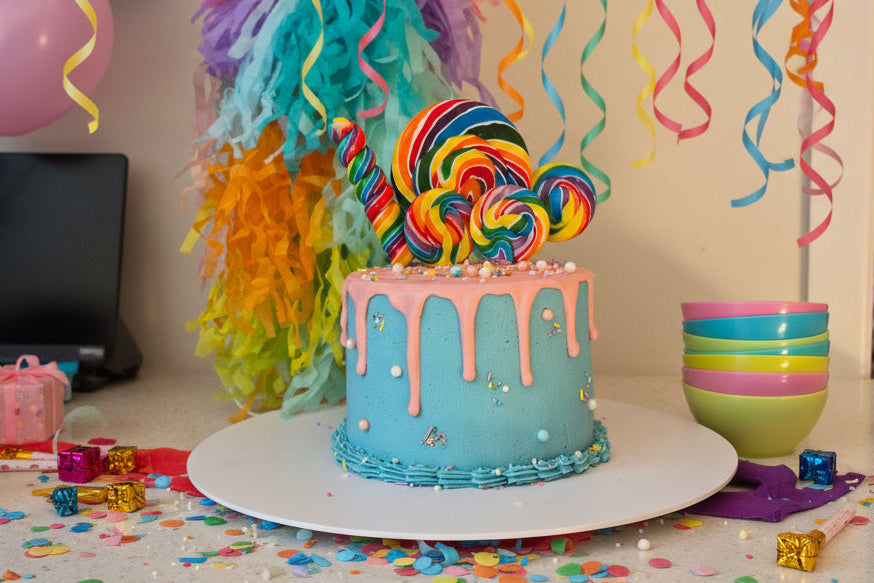 A decorated cake on a white round stand, featuring light blue frosting, pink drip icing, and sprinkles, alongside a large rainbow-striped lollipop decoration, with colorful paper shred in the background.