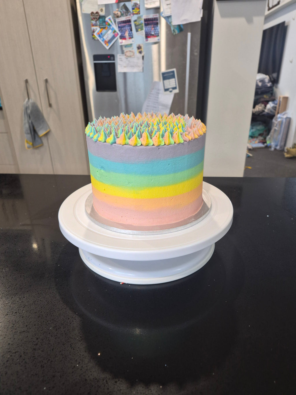 A cylindrical cake with pastel rainbow stripes in pink, yellow, green, blue, and purple, topped with yellow and green piped frosting, mounted on a white rotating cake stand.