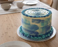 A round cake with blue and white marbled frosting and piped blue ruffle trim, displayed on a wooden surface next to a tiled wall.