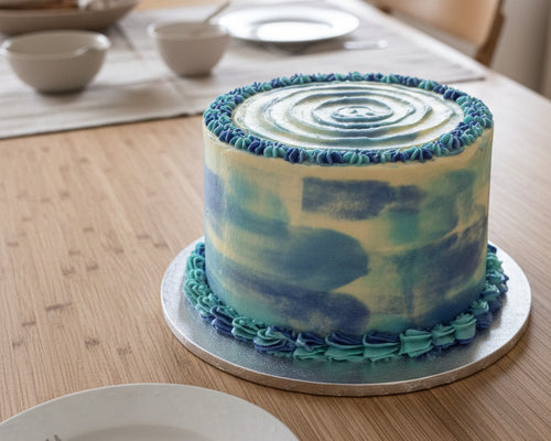 A round cake with blue and white marbled frosting and piped blue ruffle trim, displayed on a wooden surface next to a tiled wall.