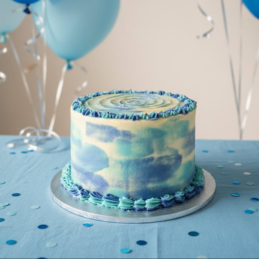 A round cake with blue and white watercolor buttercream icing and piped blue ruffle trim, displayed on a wooden surface next to a tiled wall.